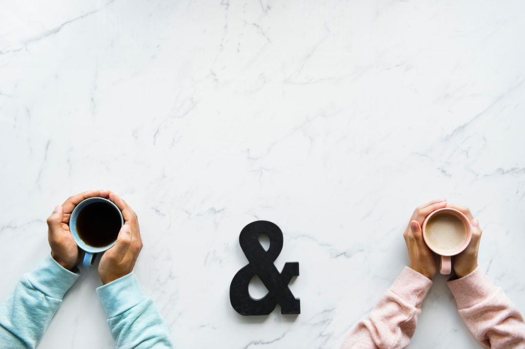 Top down view of two pairs of hands – each wrapped around a hot drink with an “&” sign in the middle.