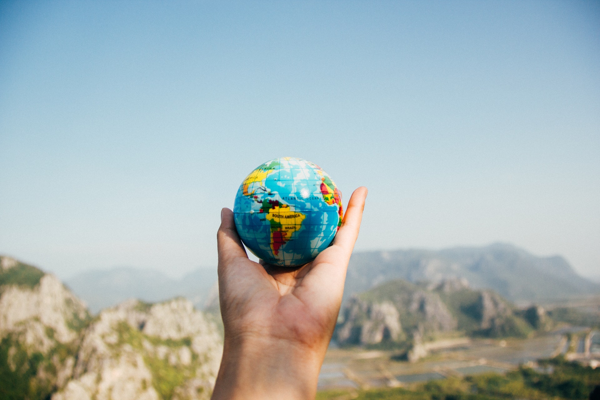 An open hand holding a palm sized world globe in the air against a dramatic mountainous backdrop.