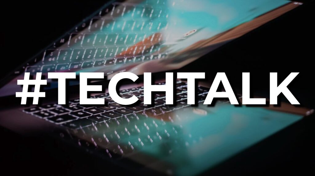 #TECHTALK hashtag in large bold white text with offset shadow over a dark photo of a half open laptop with the screen on with bright blue reflecting some of the keys